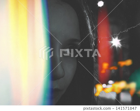 Moody cinematic portrait of a young woman partially hidden behind a rainbow prism light leak on a city street at night. Analog film aesthetic. Moody cinematic portrait of a young woman partially hidden behind a rainbow prism light leak on a city street at night. Analog film aesthetic. 141571847