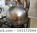 close-up shot of very dirty metal kettle sitting on a gas stove burner, showing significant signs of use with burnt stains, scratches, and discoloration on surface 141572044