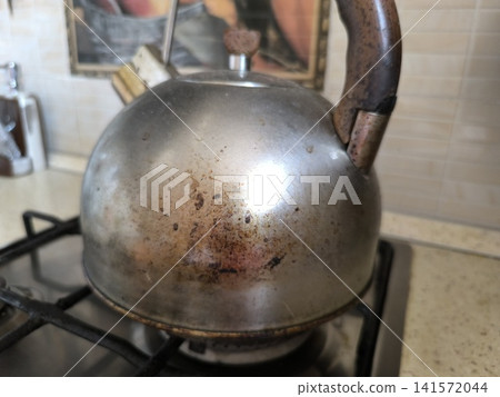 close-up shot of very dirty metal kettle sitting on a gas stove burner, showing significant signs of use with burnt stains, scratches, and discoloration on surface close-up shot of very dirty metal kettle sitting on a gas stove burner, showing significant signs of use with burnt stains, scratches, and discoloration on surface 141572044