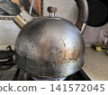 close-up shot of very dirty metal kettle sitting on a gas stove burner, showing significant signs of use with burnt stains, scratches, and discoloration on surface 141572045