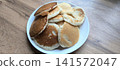 white ceramic plate piled with golden-brown fluffy homemade pancakes, captured in natural lighting on a light wood grain table surface 141572047