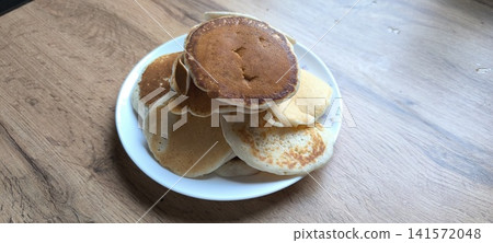 white ceramic plate piled with golden-brown fluffy homemade pancakes, captured in natural lighting on a light wood grain table surface 141572048