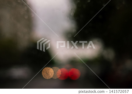 Rain drops on a car window with blurred city traffic and glowing red and yellow lights Moody urban weather scene capturing rainy gloomy autumnal day. Rain drops on a car window with blurred city traffic and glowing red and yellow lights Moody urban weather scene capturing rainy gloomy autumnal day. 141572057