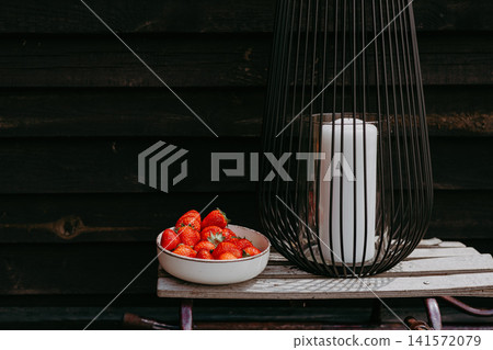 Fresh strawberries in a ceramic bowl next to a modern black lantern with white candle on rustic wooden surface outdoors. Summer picnic on the terrace. Fresh strawberries in a ceramic bowl next to a modern black lantern with white candle on rustic wooden surface outdoors. Summer picnic on the terrace. 141572079