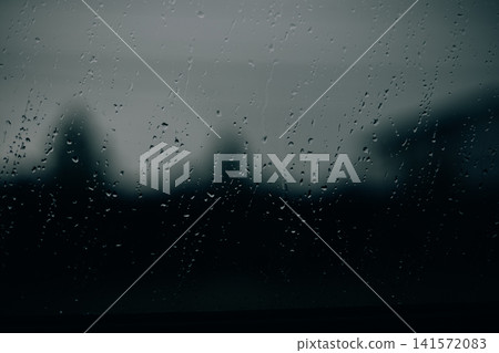 Raindrops on a window during heavy rainfall with blurred residential houses in the background. Atmospheric image representing stormy weather. Raindrops on a window during heavy rainfall with blurred residential houses in the background. Atmospheric image representing stormy weather. 141572083