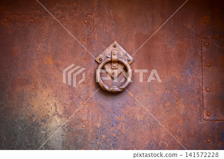 Antique metal door knocker with decorative ring mounted on a rusty corroded door surface. 141572228