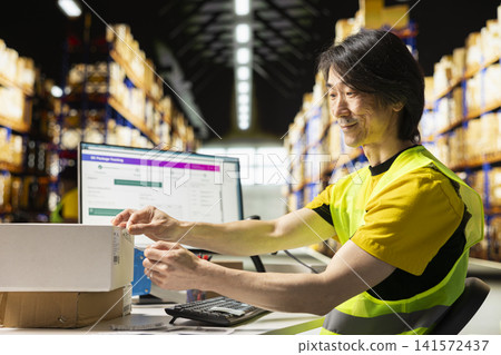 Asian male worker printing tracking labels and applying on packages, processing orders and managing labeling for easy shipment tracking on pc software. Fulfillment center full of racks. Asian male worker printing tracking labels and applying on packages, processing orders and managing labeling for easy shipment tracking on pc software. Fulfillment center full of racks. 141572437