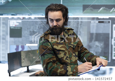 Military intelligence officer writes enemy coordinates on clipboard in command center. Army specialist records target data and defensive operation information from surveillance screens Military intelligence officer writes enemy coordinates on clipboard in command center. Army specialist records target data and defensive operation information from surveillance screens 141572457