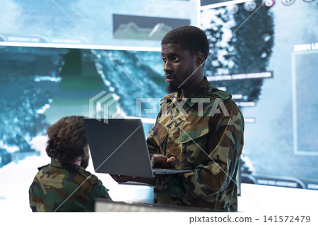 Military intelligence specialist using laptop to analyze defense systems data. African american man checking surveillance target feeds in war room with notebook 141572479