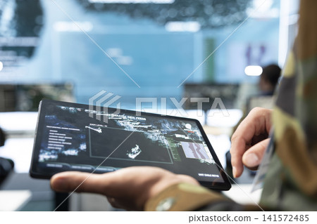 Close up of military specialist using tablet displaying aerial tactical map. Army operator selects targets on device touchscreen while monitoring command center surveillance 141572485