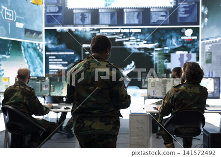 Team of military officers monitoring surveillance displays across operations room. Army supervisors overseeing operators tracking satellite maps and target information Team of military officers monitoring surveillance displays across operations room. Army supervisors overseeing operators tracking satellite maps and target information 141572492