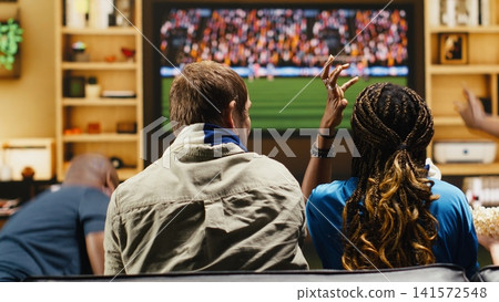 Football supporters at home argue over referee decision, shouting and raising hands during tense match. Soccer fans show frustration through frantic gestures, reacting to dramatic live game on TV Football supporters at home argue over referee decision, shouting and raising hands during tense match. Soccer fans show frustration through frantic gestures, reacting to dramatic live game on TV 141572548
