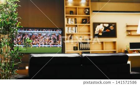 Soccer game broadcasted on TV in empty apartment living room before fans arrive. Cozy sofa, plant and television set screen creating comfortable home viewing setup for sports event Soccer game broadcasted on TV in empty apartment living room before fans arrive. Cozy sofa, plant and television set screen creating comfortable home viewing setup for sports event 141572586