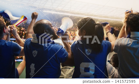 Football fans in blue jerseys sounding horns and megaphone inside sports arena during match. Soccer supporters blowing vuvuzelas and waving scarves in stadium stands Football fans in blue jerseys sounding horns and megaphone inside sports arena during match. Soccer supporters blowing vuvuzelas and waving scarves in stadium stands 141572595