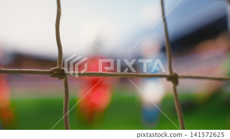 Selective focus of soccer ball crossing line of goal frame on pitch, goal shot hitting net. Red soccer players of winning team cheering near defeated opponents, fallen goalie in blurred motion. Selective focus of soccer ball crossing line of goal frame on pitch, goal shot hitting net. Red soccer players of winning team cheering near defeated opponents, fallen goalie in blurred motion. 141572625