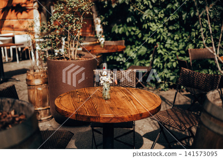 Cozy outdoor cafe terrace featuring a round wooden table, woven chairs and a vase of wildflowers. Rustic textures create an inviting atmosphere Cozy outdoor cafe terrace featuring a round wooden table, woven chairs and a vase of wildflowers. Rustic textures create an inviting atmosphere 141573095