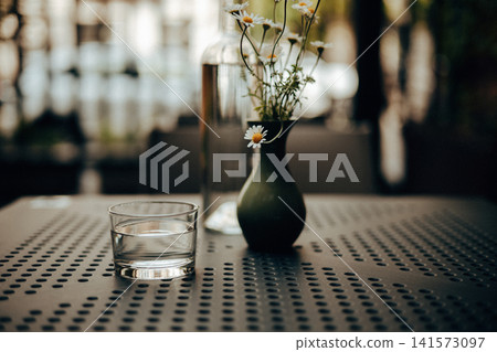 Minimalist still life featuring a glass of water, a small vase with delicate chamomile flowers, a clear bottle placed on a perforated metal table. A calm, cozy cafe terrace atmosphere. Modern interior Minimalist still life featuring a glass of water, a small vase with delicate chamomile flowers, a clear bottle placed on a perforated metal table. A calm, cozy cafe terrace atmosphere. Modern interior 141573097
