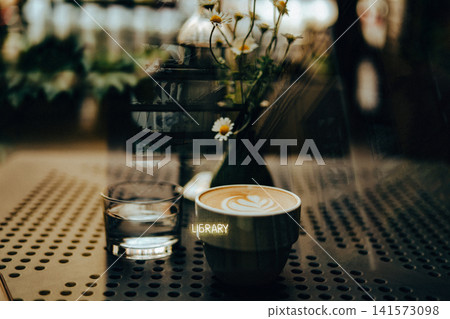 A crafted latte with delicate heart-shaped latte art sits beside a glass of water on a perforated cafe table, soft reflections on the window create layered visual depth. Wild daisies in a small vase. A crafted latte with delicate heart-shaped latte art sits beside a glass of water on a perforated cafe table, soft reflections on the window create layered visual depth. Wild daisies in a small vase. 141573098