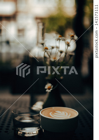 A beautifully crafted latte with heart-shaped latte art sits on a perforated metal cafe table beside a glass of water and delicate chamomile flowers. 141573111