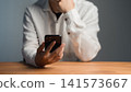 Modern smartphone technology concept. Serious adult man thinking while holding a mobile phone in a home office. Online shopping, customer service and payment. 141573667