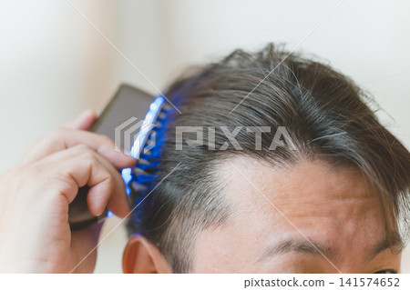 A man in his 40s with graying hair is using a blue LED brush on his head. A man in his 40s with graying hair is using a blue LED brush on his head. 141574652