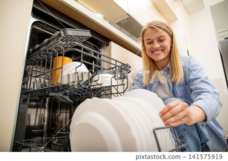 Dishwasher. Woman smiles while unloading clean dishes from dishwasher in stylish kitchen Dishwasher. Woman smiles while unloading clean dishes from dishwasher in stylish kitchen 141575919
