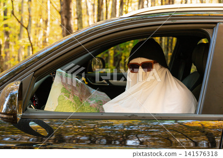 Person wearing ghost costume driving car while holding paper map, searching for direction and navigating route Person wearing ghost costume driving car while holding paper map, searching for direction and navigating route 141576318