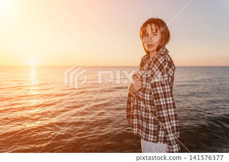 Sunset woman beach standing near the peaceful ocean during golden hour wearing a casual plaid shirt with copy space 141576377