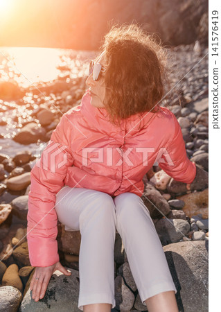 Woman beach sunset sitting on a rocky shore wearing a pink puffer jacket during golden hour for travel concept Woman beach sunset sitting on a rocky shore wearing a pink puffer jacket during golden hour for travel concept 141576419