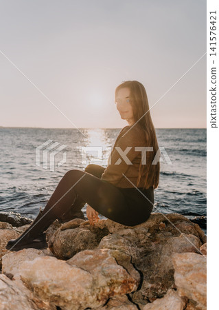Woman sunset ocean sitting on rocky shore while looking back over shoulder toward golden horizon at dusk Woman sunset ocean sitting on rocky shore while looking back over shoulder toward golden horizon at dusk 141576421