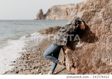 Woman beach rock leaning on a large boulder while barefoot on a pebble seashore with coastal cliffs in the background Woman beach rock leaning on a large boulder while barefoot on a pebble seashore with coastal cliffs in the background 141576422