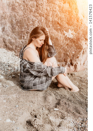 Woman beach plaid sitting on sandy shore by rocky cliff while looking down thoughtfully in warm sunlight during summer vacation 141576423