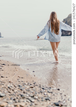 Woman beach ocean walking along the shore at sunset wearing an oversized blue shirt enjoying a summer vacation day Woman beach ocean walking along the shore at sunset wearing an oversized blue shirt enjoying a summer vacation day 141576427