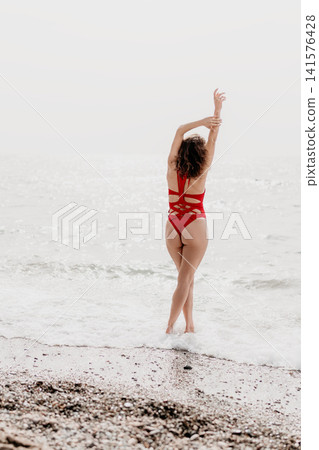 Beach swimsuit woman wearing red swimwear with arms raised on a hazy shoreline enjoying summer vacation relaxation with copy space 141576428