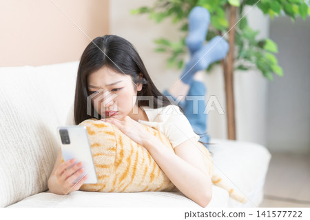 一位年輕女子麵朝下躺在沙發上,神情焦慮地看著她的智慧型手機。 一位年輕女子麵朝下躺在沙發上,神情焦慮地看著她的智慧型手機。 141577722