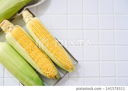 玉米:帶皮的生玉米棒,放在白色磁磚上。 玉米:帶皮的生玉米棒,放在白色磁磚上。 141578311