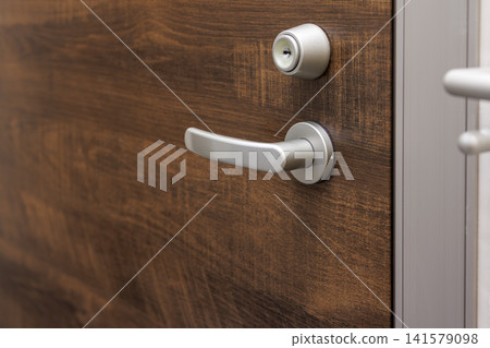 A silver door lever handle and lock are attached to a dark brown wood-grain door. A silver door lever handle and lock are attached to a dark brown wood-grain door. 141579098