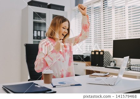 Confident and Composed: A professional businesswoman exudes confidence and composure as she works on her laptop, surrounded by the vibrant energy of a modern office space. Confident and Composed: A professional businesswoman exudes confidence and composure as she works on her laptop, surrounded by the vibrant energy of a modern office space. 141579468