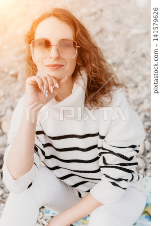 Woman beach fashion posing on a pebble shore in a striped sweater and sunglasses for a relaxed summer portrait Woman beach fashion posing on a pebble shore in a striped sweater and sunglasses for a relaxed summer portrait 141579626