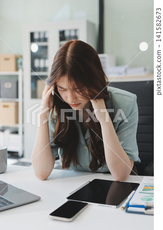 Asian businesswoman having a stressful phone call, holding a pen, and looking troubled while working on her laptop with financial documents and a calculator on her office desk Asian businesswoman having a stressful phone call, holding a pen, and looking troubled while working on her laptop with financial documents and a calculator on her office desk 141582673