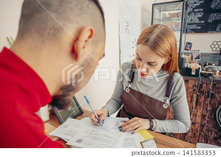 A young Caucasian woman with red hair assists a man at a cafe. She wears a brown apron and writes on a paper. The cafe has a cozy atmosphere with coffee equipment visible. A young Caucasian woman with red hair assists a man at a cafe. She wears a brown apron and writes on a paper. The cafe has a cozy atmosphere with coffee equipment visible. 141583353