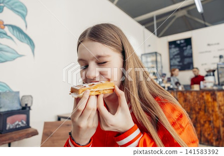 A young Caucasian girl with long brown hair enjoys a pastry in a modern cafe. The background features a barista and a cozy atmosphere. A young Caucasian girl with long brown hair enjoys a pastry in a modern cafe. The background features a barista and a cozy atmosphere. 141583392