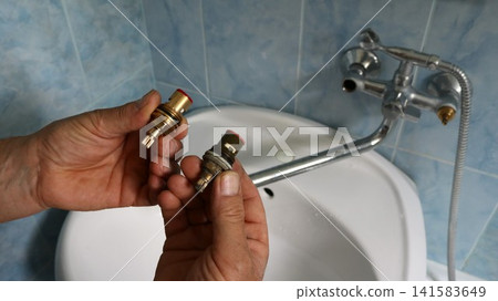 A man's hands hold two faucet valves: a new, shiny one and an old, worn one. In the background, a disassembled wall-mounted faucet is visible above a white sink in a bathroom tiled with blue tiles. A man's hands hold two faucet valves: a new, shiny one and an old, worn one. In the background, a disassembled wall-mounted faucet is visible above a white sink in a bathroom tiled with blue tiles. 141583649