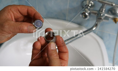 A man's hands hold a disassembled faucet aerator: a round mesh filter and a metal sleeve attachment. The process takes place over a white sink in a bathroom with blue tile on the wall. A man's hands hold a disassembled faucet aerator: a round mesh filter and a metal sleeve attachment. The process takes place over a white sink in a bathroom with blue tile on the wall. 141584270