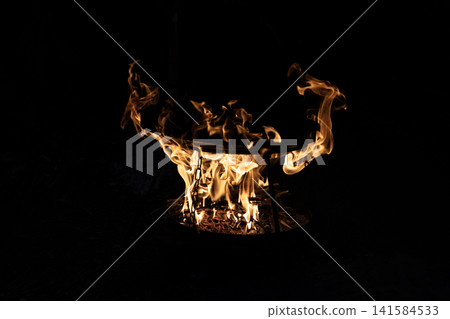 Bright burning wood fire inside a metal round grill bowl with glowing flames in the dark night. Close up view of outdoor campfire cooking setup with hot orange fire tongues shaping abstract patterns. Bright burning wood fire inside a metal round grill bowl with glowing flames in the dark night. Close up view of outdoor campfire cooking setup with hot orange fire tongues shaping abstract patterns. 141584533