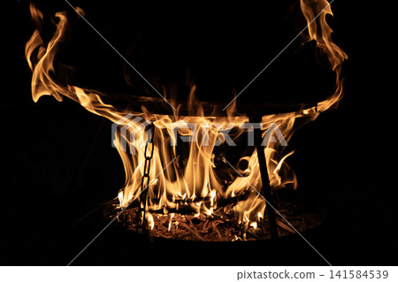 Bright burning wood fire inside a metal round grill bowl with glowing flames in the dark night. Close up view of outdoor campfire cooking setup with hot orange fire tongues shaping abstract patterns. 141584539