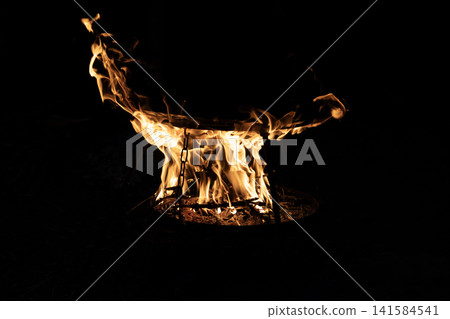 Bright burning wood fire inside a metal round grill bowl with glowing flames in the dark night. Close up view of outdoor campfire cooking setup with hot orange fire tongues shaping abstract patterns. Bright burning wood fire inside a metal round grill bowl with glowing flames in the dark night. Close up view of outdoor campfire cooking setup with hot orange fire tongues shaping abstract patterns. 141584541
