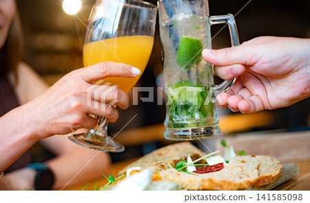 Girl Toasts With Friend At Bar Table Using Beer And Mojito Glasses Girl Toasts With Friend At Bar Table Using Beer And Mojito Glasses 141585098