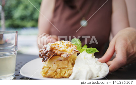 Girl Enjoys Apple Charlotte With Cream And Mint, Sipping Cold Lemonade At A Breezy Outdoor Cafe Girl Enjoys Apple Charlotte With Cream And Mint, Sipping Cold Lemonade At A Breezy Outdoor Cafe 141585107