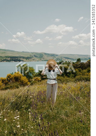 girl holds a hat covering her face on a sunny day in a grassy meadow with a view of the Tbilisi Sea girl holds a hat covering her face on a sunny day in a grassy meadow with a view of the Tbilisi Sea 141585352